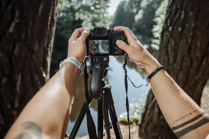Hände halten eine Kamera auf einem Stativ, im Hintergrund ein Waldsee